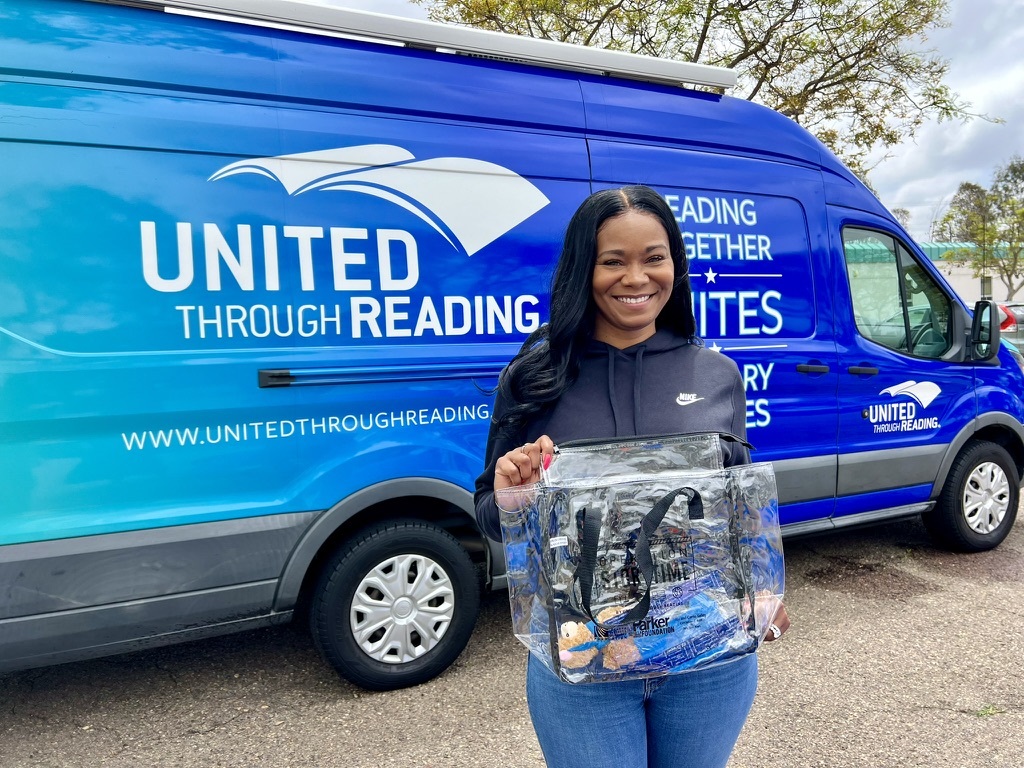 woman holds UTR storytime kit in front of United Through Reading Mobile Story Station