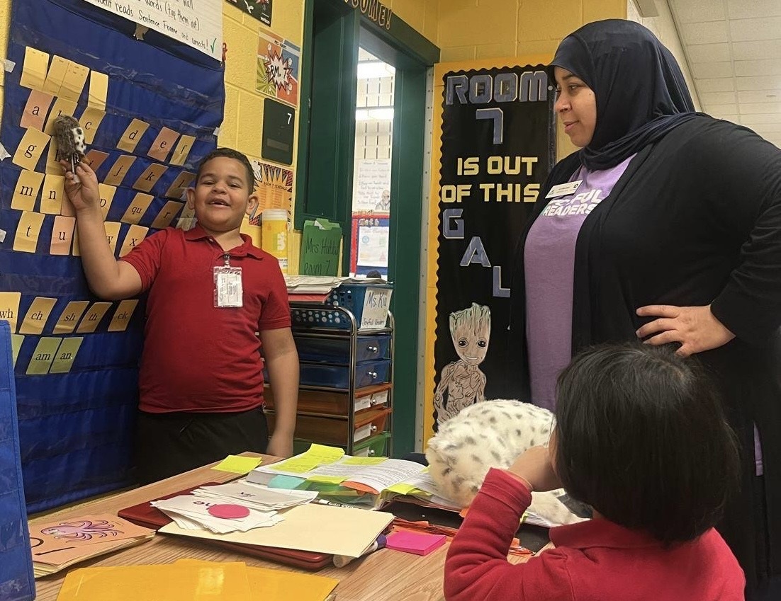 Joyful Readers male student recites letters from board with female Muslim teacher looking on